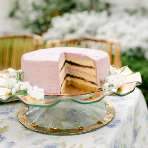 Pink layered cake on a glass 24k gold Ruffle cake plate with white flowers on a floral tablecloth.
