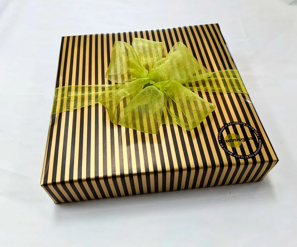 An example of Annieglass's gift-wrapped box with black and gold stripes, tied with a lime green ribbon and adorned with a gold dot pattern. An Annieglass sticker is visible on the box.