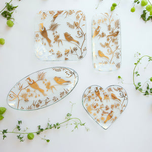 Decorative glass plates with 24k gold bird designs on a white surface with greenery
