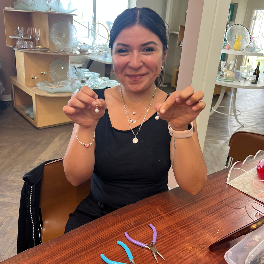 Woman holding up two pearl earrings with jewelry-making tools on a table.