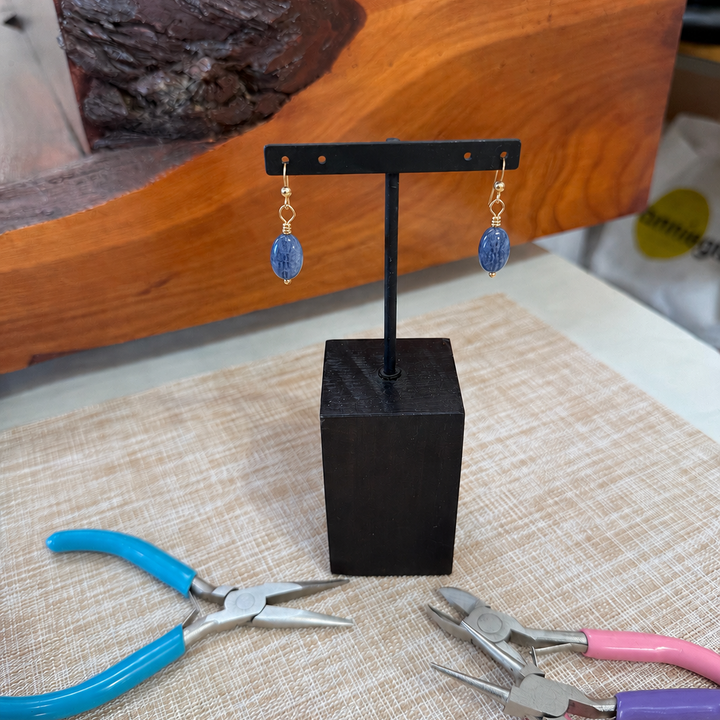 Kyanite wire earrings hanging on a black stand on a tan placemat with jewelry tools beside it.