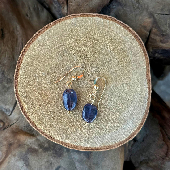 A pair of gold-tone dangle earrings with dark blue oval kyanite gemstones, displayed on a round wooden slice against a rustic wood bark background.