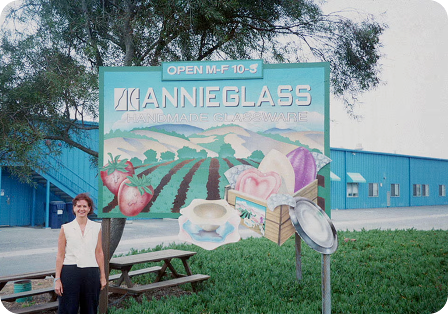 Annie in 1996 standing in front of Annieglass sign with scenic illustration and blue building in background.