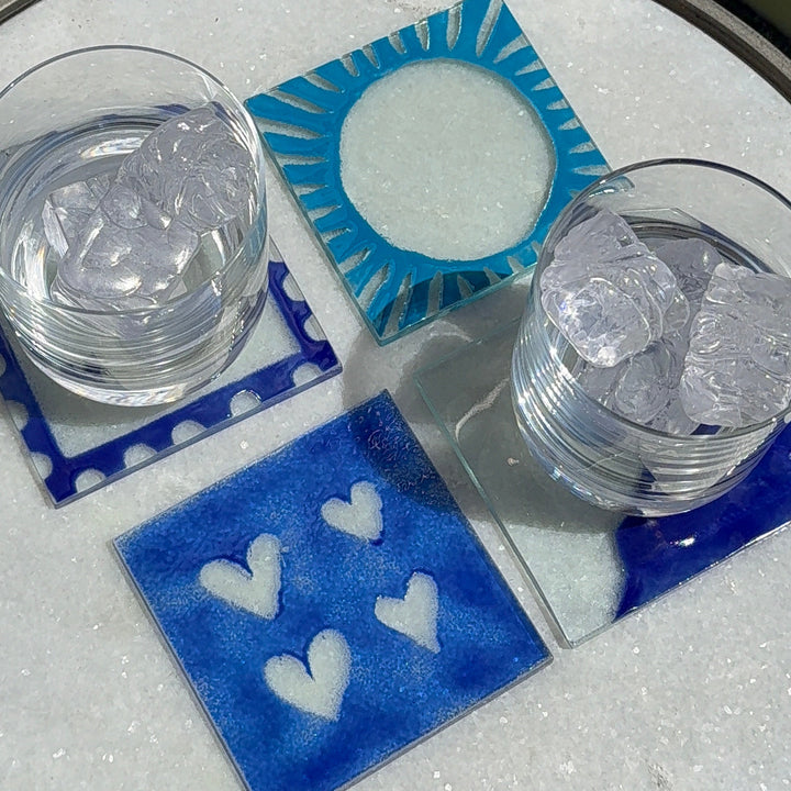 4 glass coasters with blue designs; one with hearts, one a sunburst, one a wave, and one a polkadot border. 2 glasses of ice water on two coasters.
