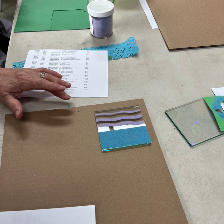 Hand reaching over a painted square glass coaster on cardboard surface with other materials on a table.
